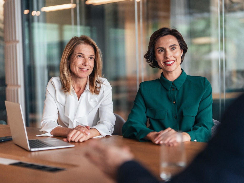 Two smiling women in business attire seated at a conference table with a laptop.