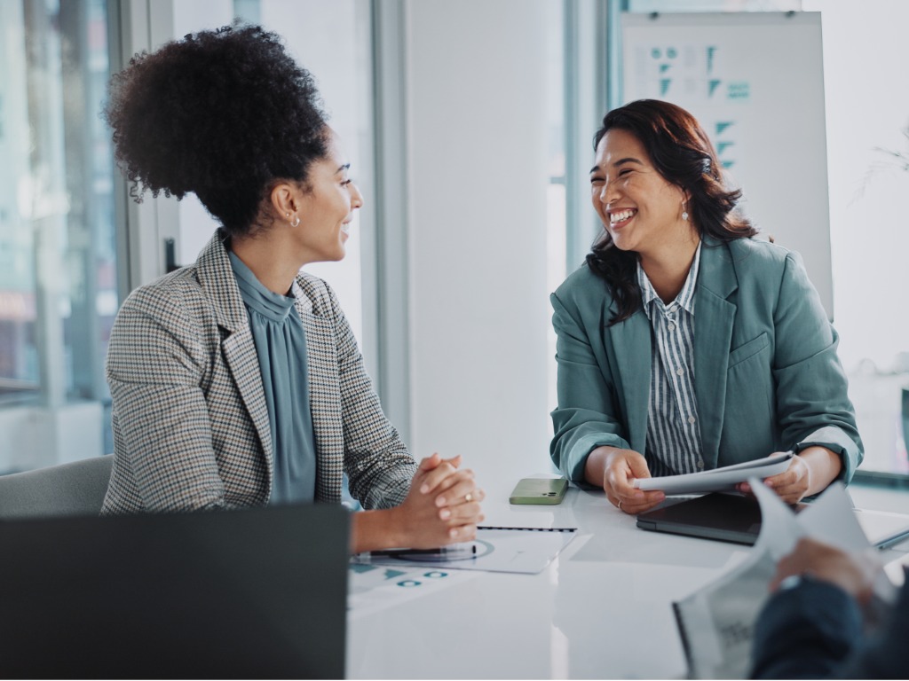 Two professional women smiling and talking at a table in a modern office meeting.