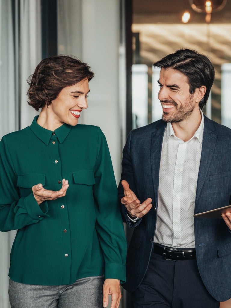 Professional man and woman in business attire discussing work, smiling and gesturing.